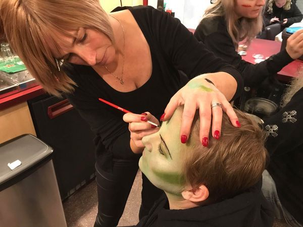 Woman applying green face paint to a child's face with a brush. Indoors.