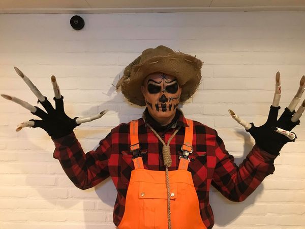 Scarecrow costume with skeletal hands, orange overalls, red plaid shirt, and straw hat.