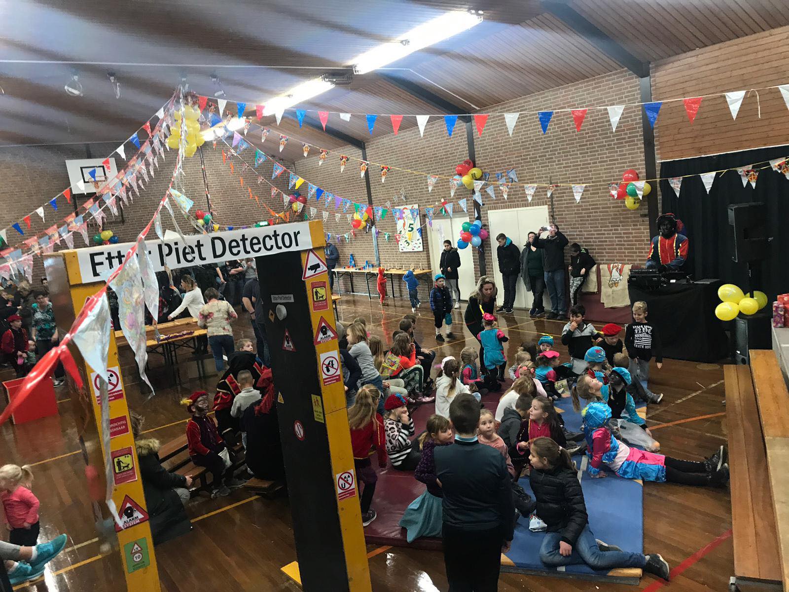 School gymnasium decorated for an event, with children and adults gathered around activity stations and a stage.