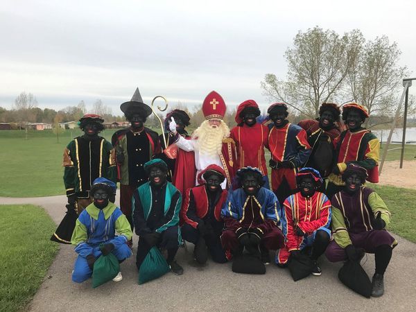 Sinterklaas with helpers in blackface costumes, posing outside near a lake.