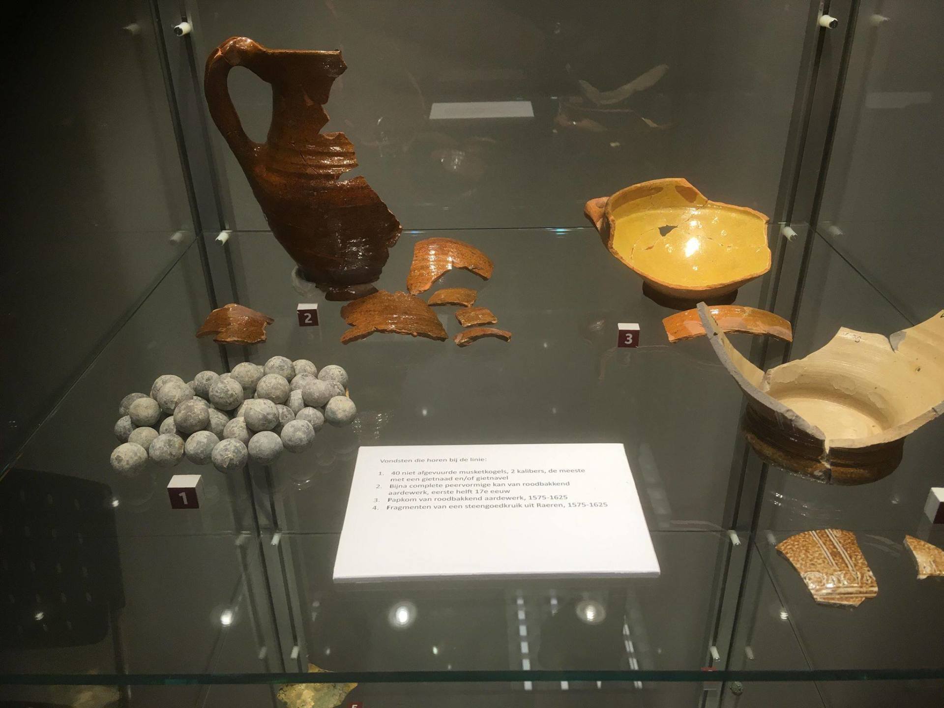 Display case of pottery fragments, a pitcher, and round projectiles. Brown, yellow, and white pottery.