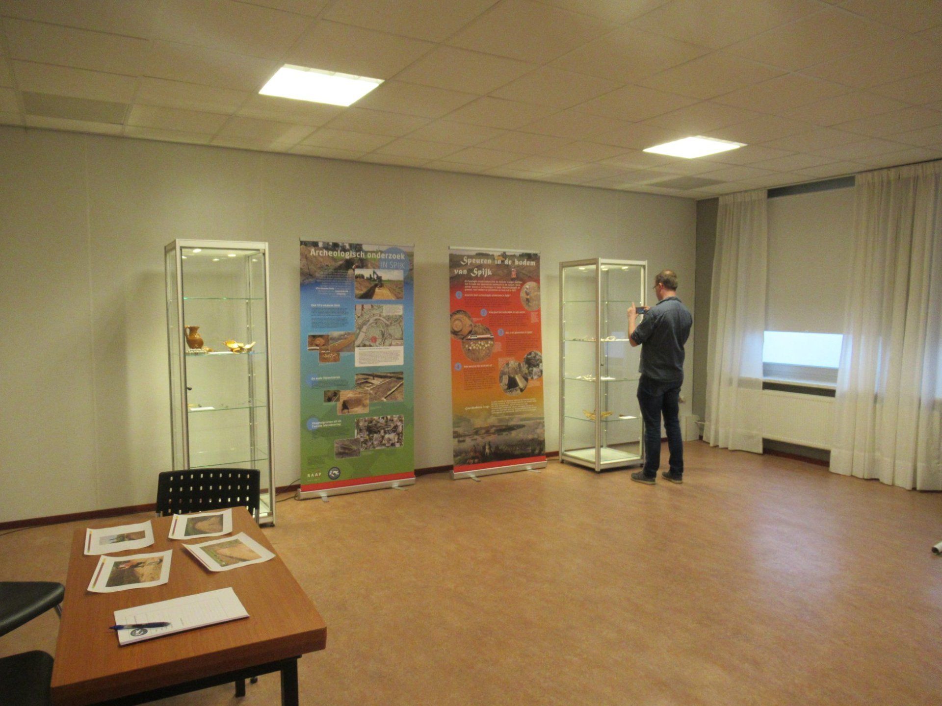 A man photographs an exhibit with display cases and posters in a room with a table and window.