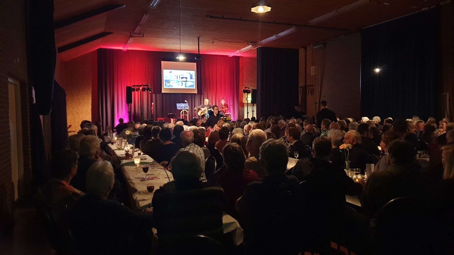 Audience seated at tables facing a stage with a presentation screen. Red lighting illuminates the stage.