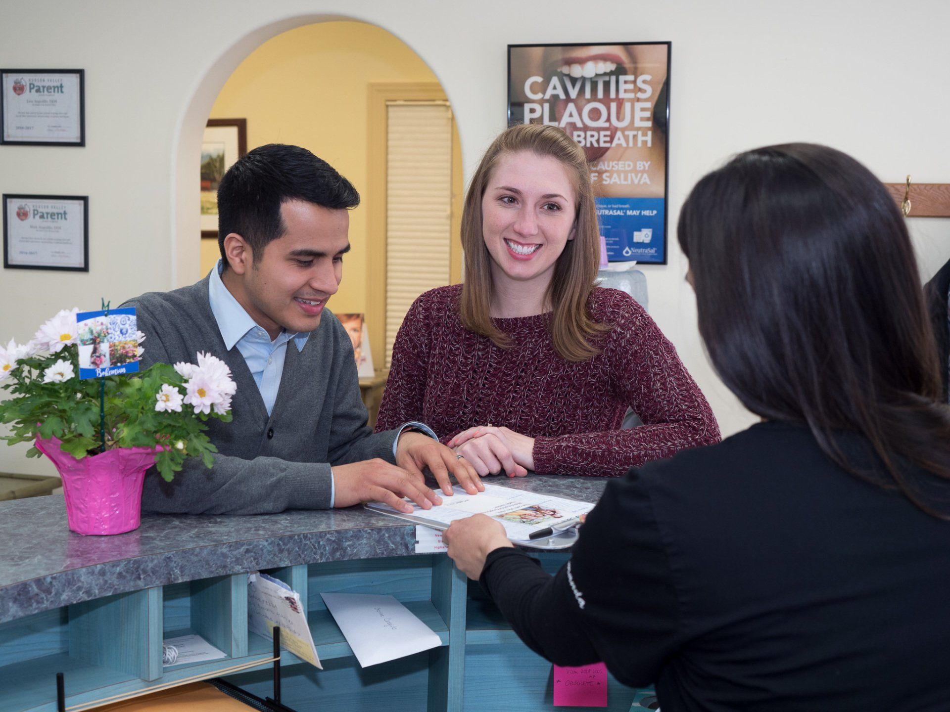 A man and woman are sitting at a counter in a dental office talking to a woman.