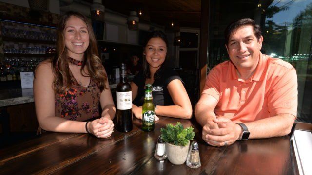 Three people are sitting at a table with a bottle of wine and a bottle of beer.