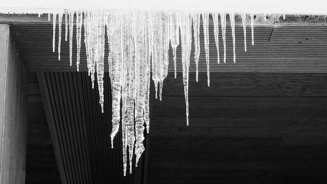 Black and white photo of icicles hanging from the underside of a concrete overpass, winter scene.