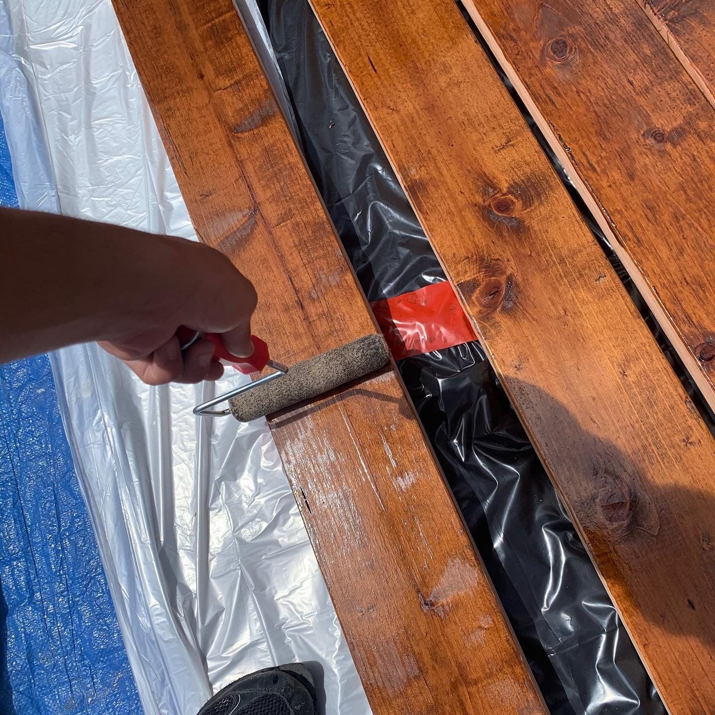 Person using a paint roller to stain wooden planks outdoors.