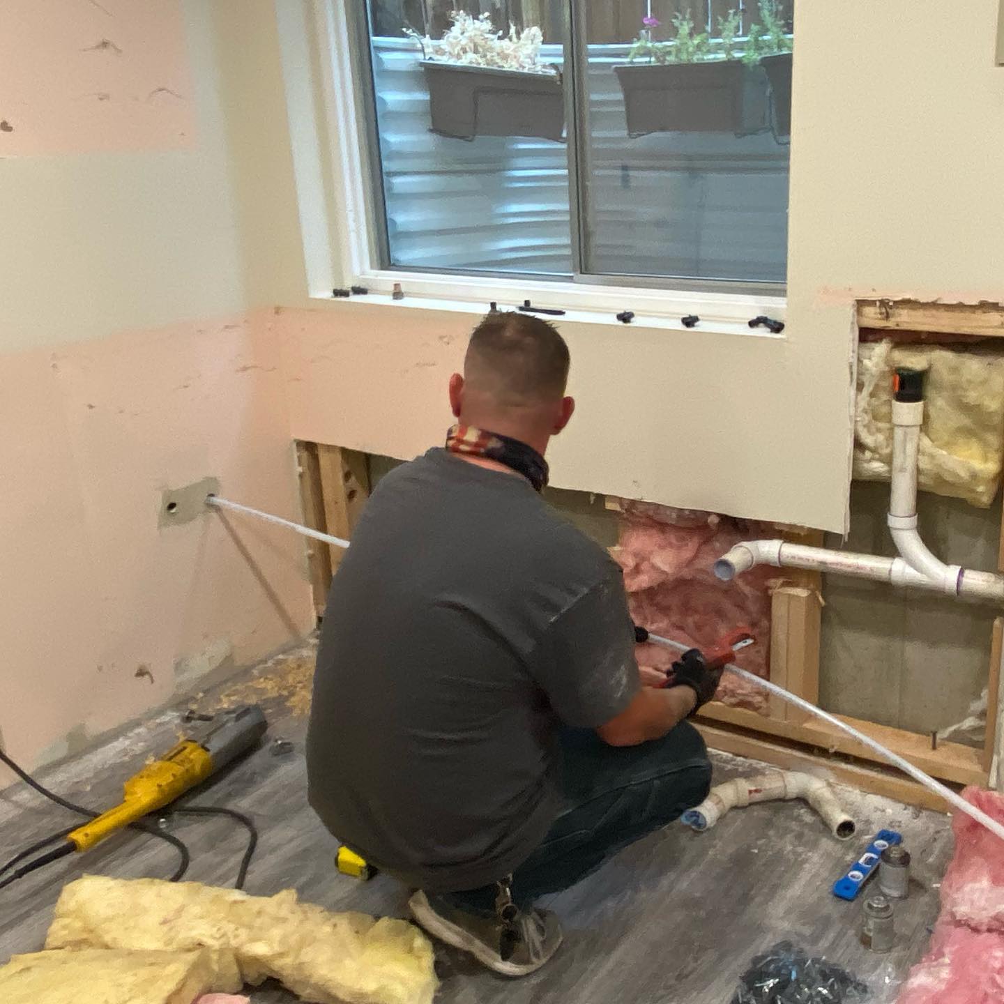 Plumber repairs pipes in a room with exposed wall studs.  A grinder sits on the floor.