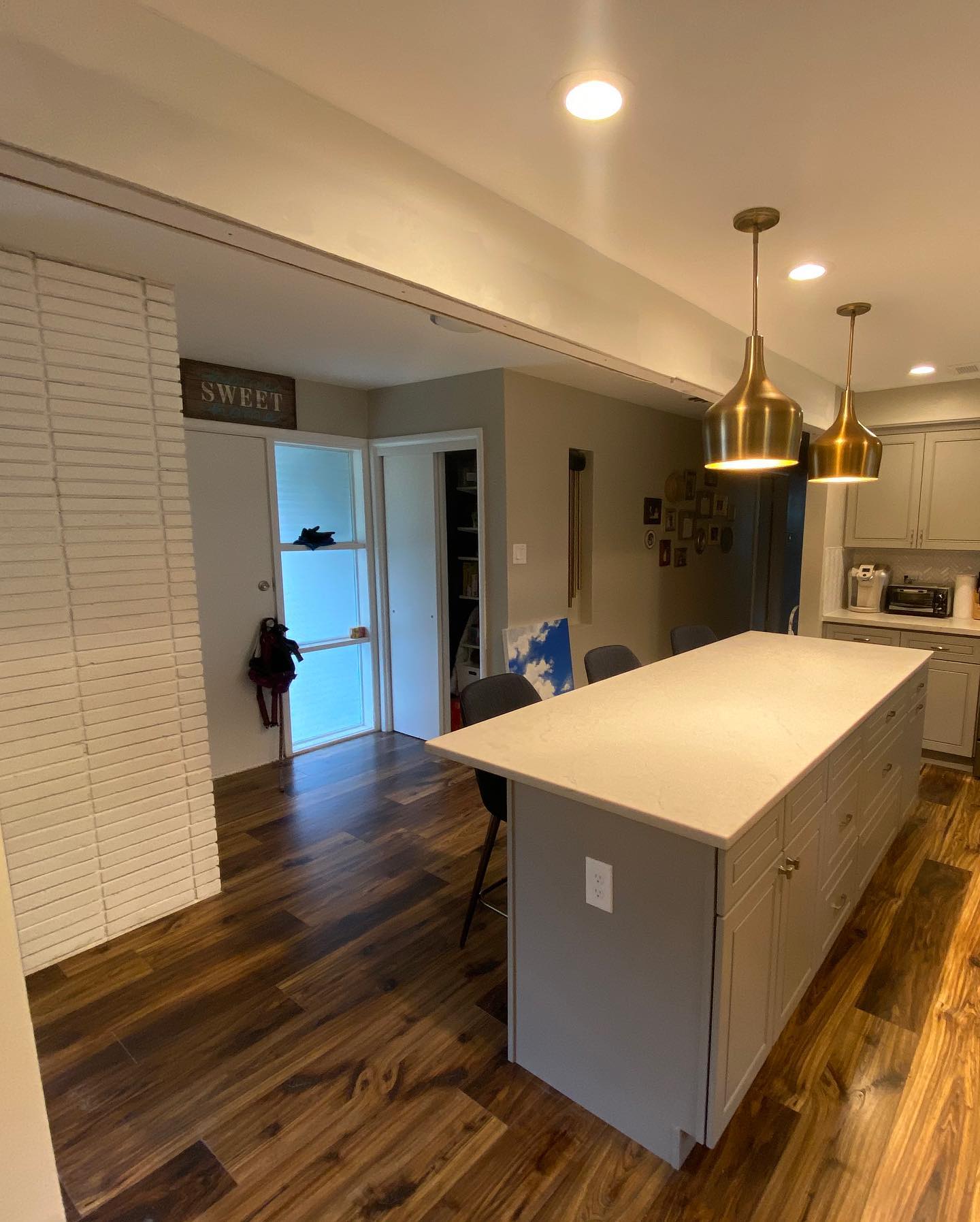 Kitchen with an island, cabinets, and wood flooring. Features a white brick wall and brass pendant lights.