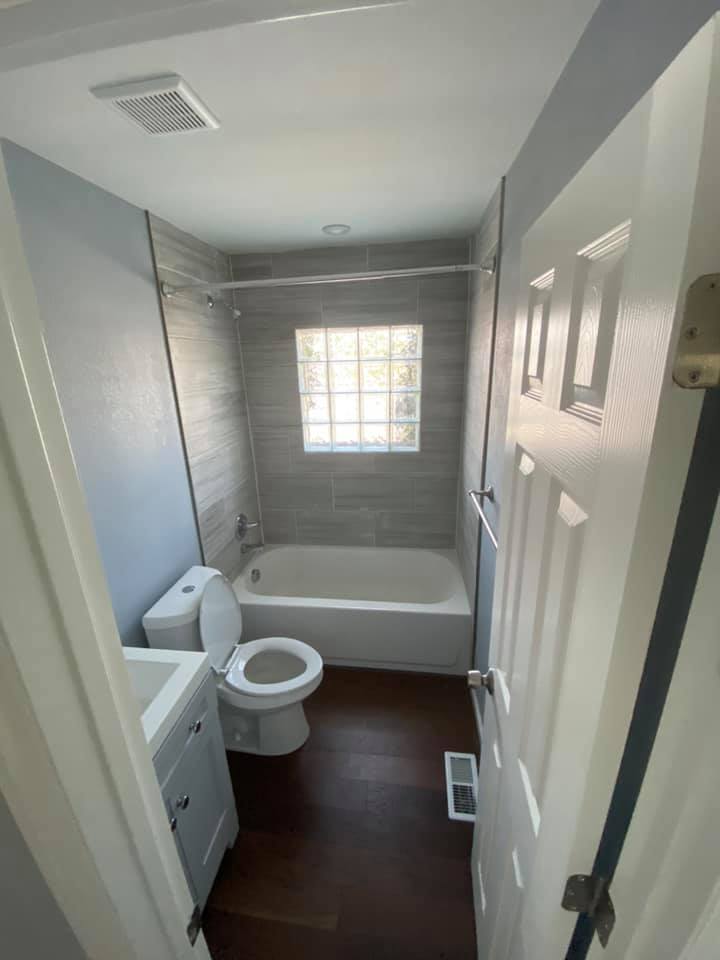 Bathroom with gray walls, wood floor, white toilet, tub, and cabinet. Window with glass blocks.