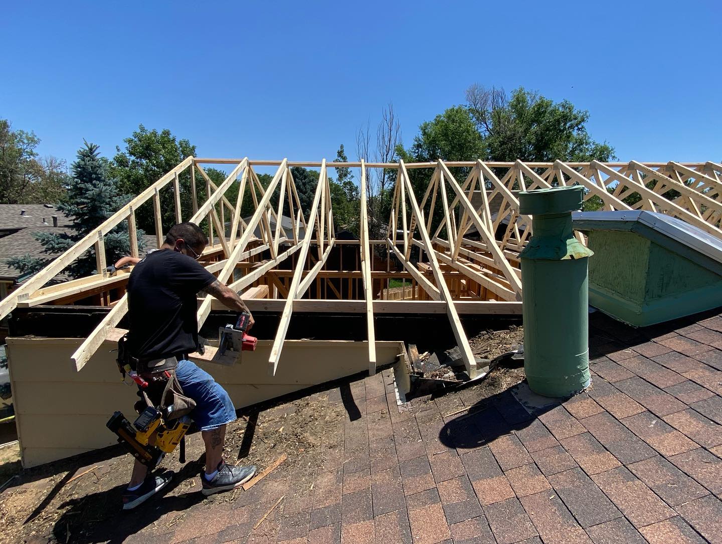 Worker on a roof installing wooden trusses beside a green vent pipe under a blue sky