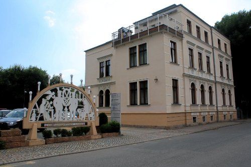 Beiges Gebäude mit dekorativem Tor davor, gepflasterter Straße und Bäumen.