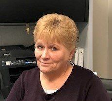 Person smiling in an office, wearing a dark top and sitting beside a printer.