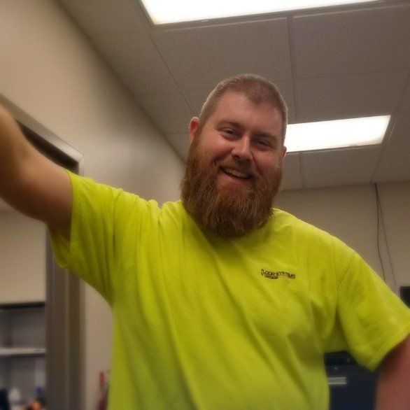 Smiling man in a neon yellow shirt raises his arm indoors under fluorescent lights.