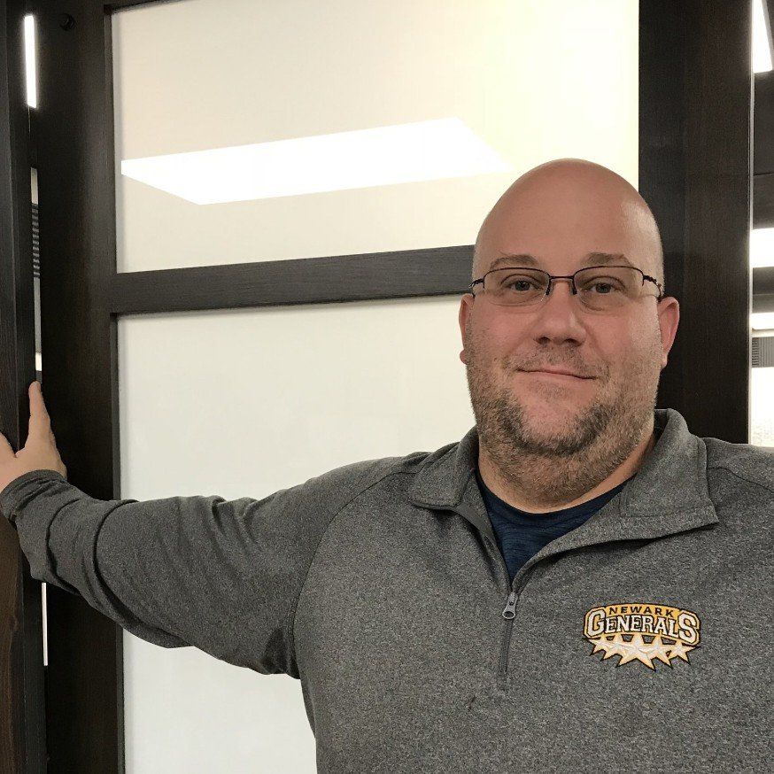 Man standing by a doorway, wearing glasses and a gray pullover, smiling slightly.