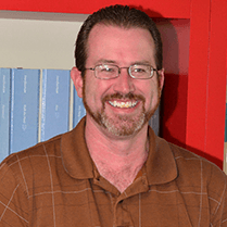 Smiling man in glasses and brown shirt standing in front of red shelving and blue binders