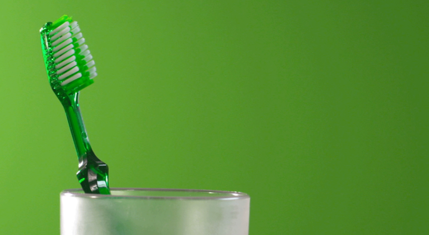 A green toothbrush is sitting in a glass on a green background.