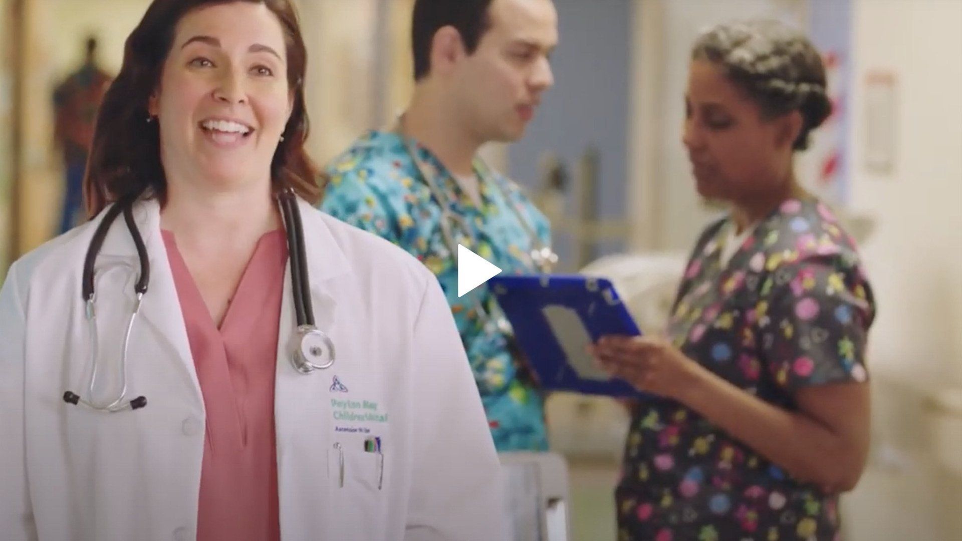 A female doctor with a stethoscope around her neck is smiling in a hospital hallway.