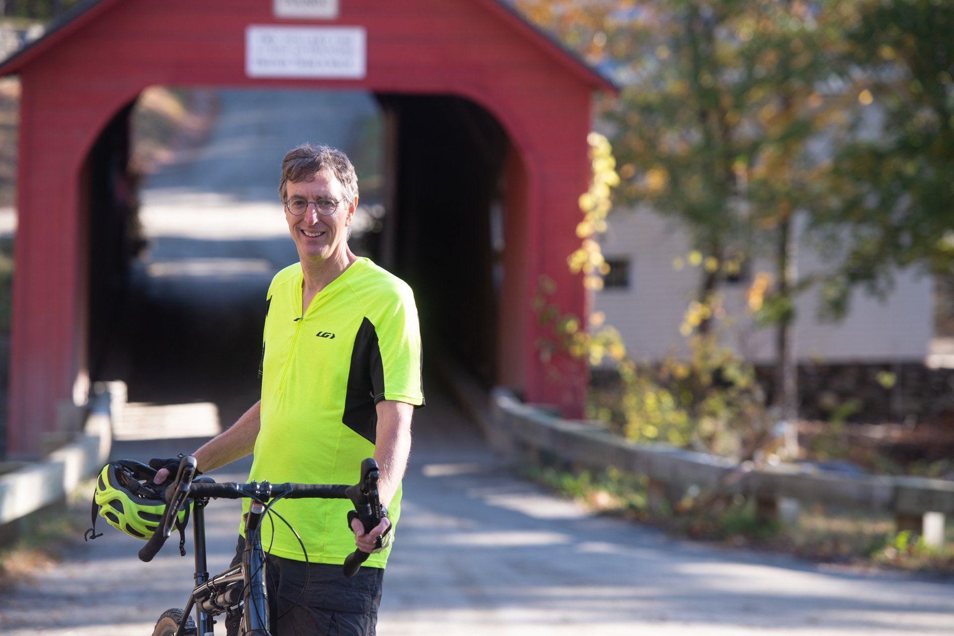 Greg Hessel in a neon yellow biking jersey standing beside a bike near a red covered bridge.