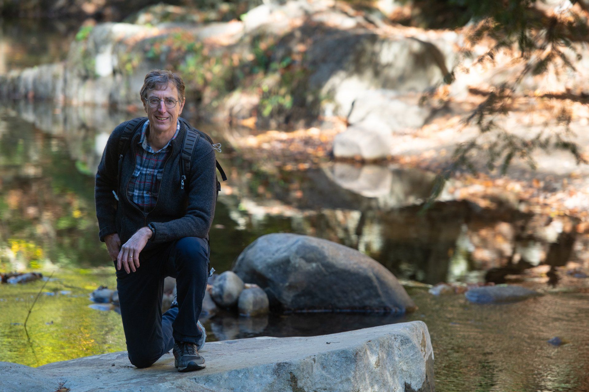 Greg Hessel sitting on a rock by a shallow creek in a wooded area, wearing a dark jacket and pants.