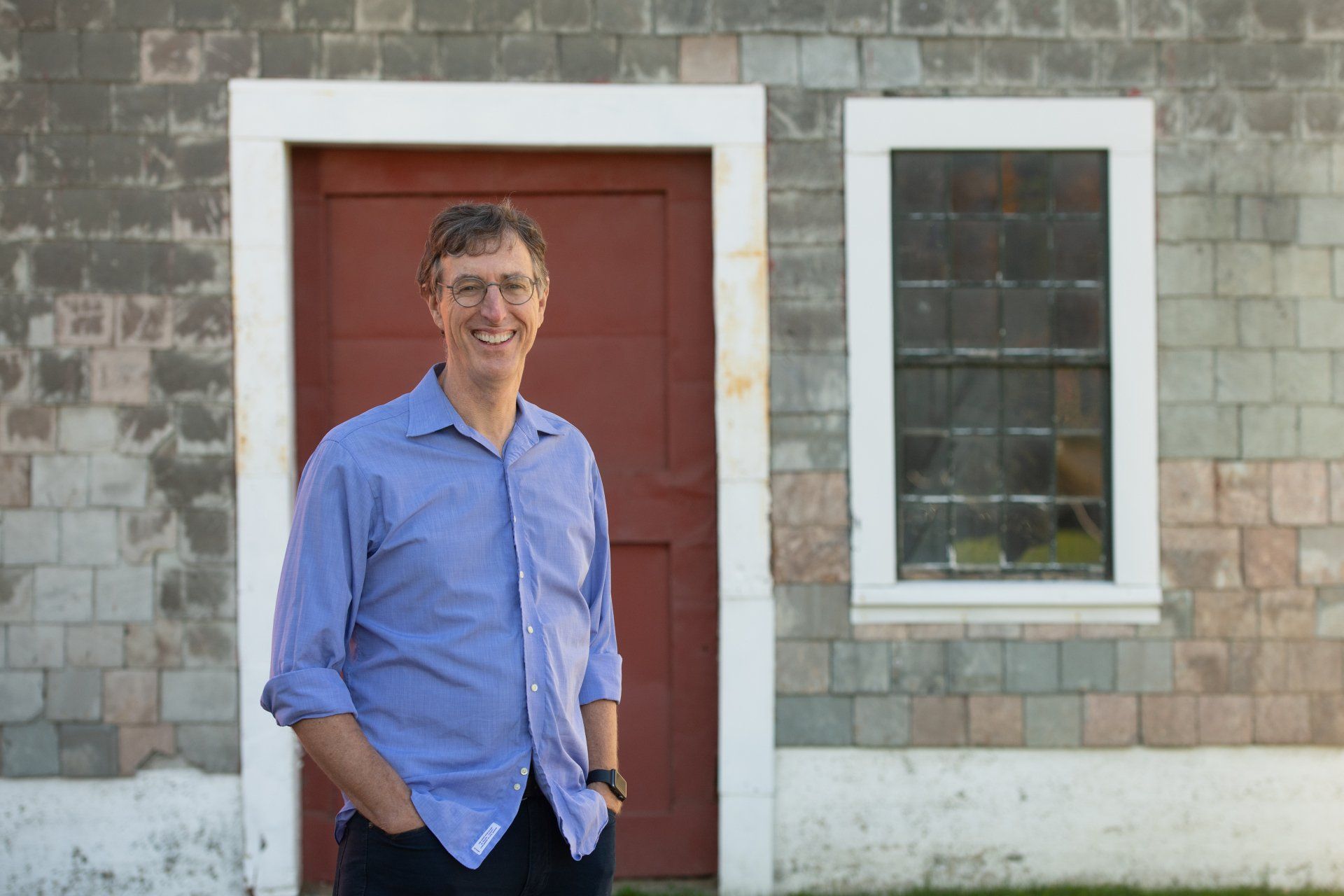 Greg Hessel in a blue shirt standing by a red door on a stone building wall