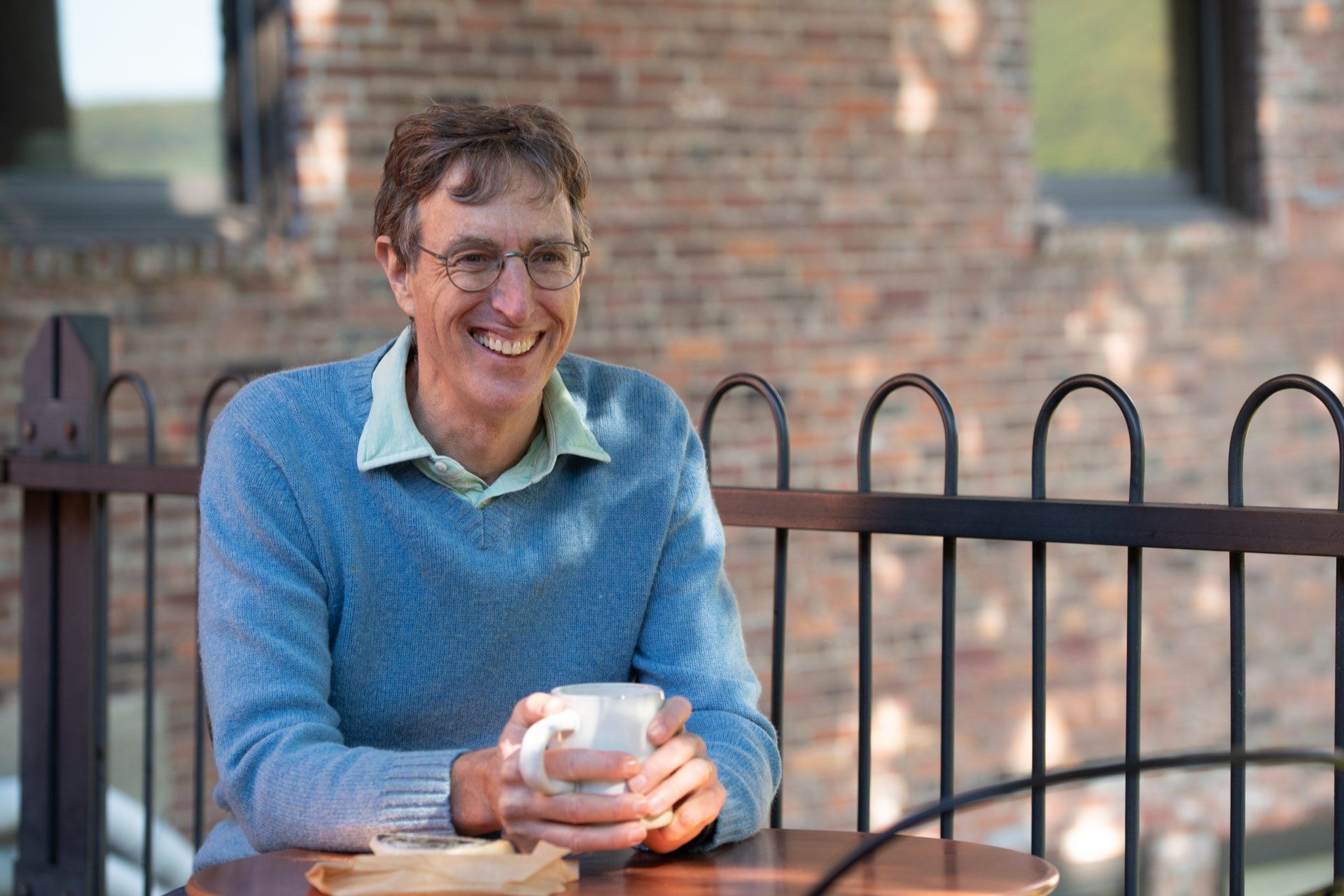Greg Hessel is smiling in a blue sweater, holding a mug at an outdoor table with a brick wall behind.