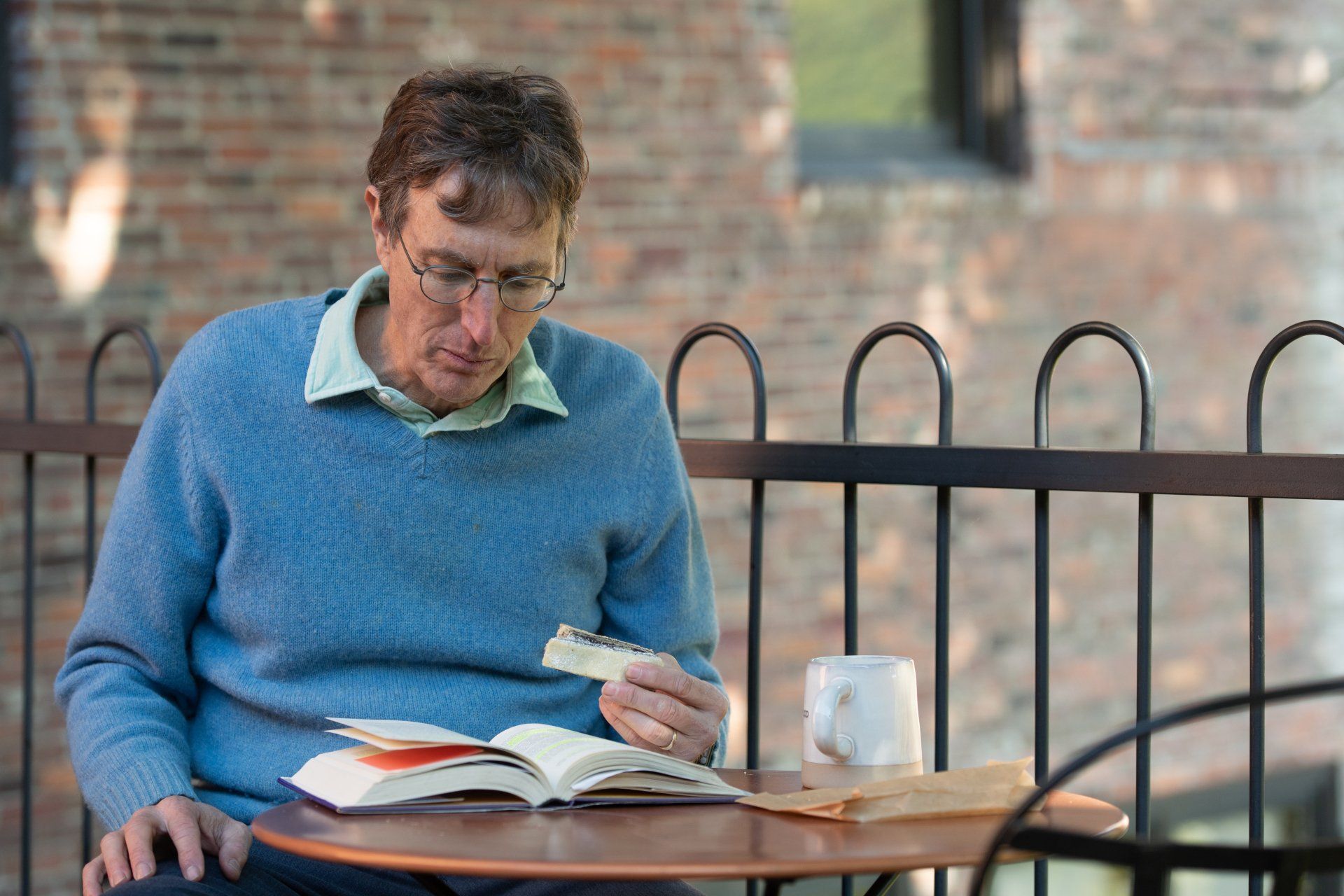 Greg Hessel in a blue sweater reading a book at an outdoor café table with a mug beside him