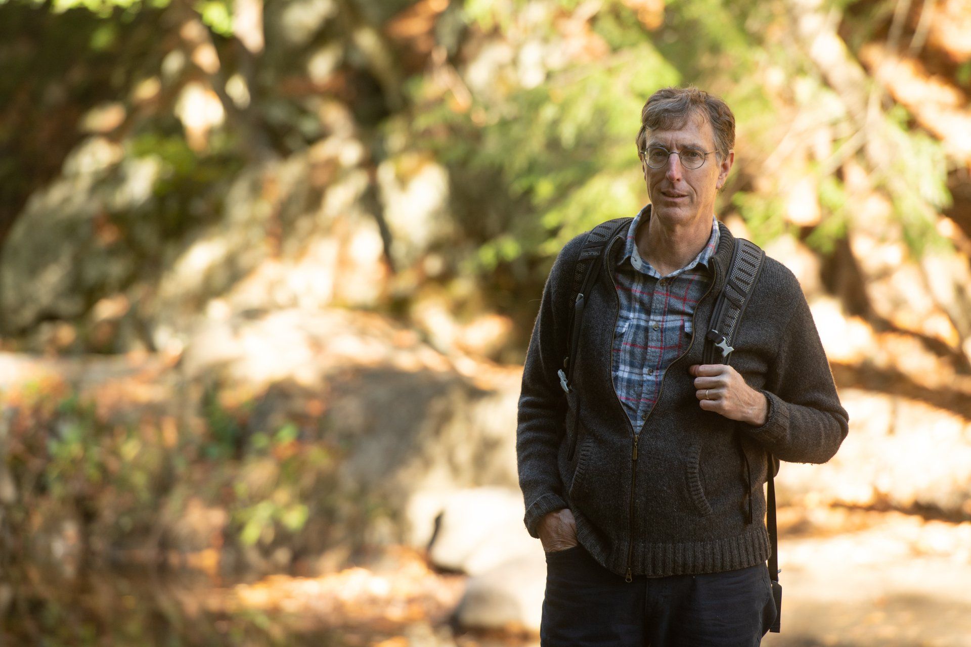Greg Hessel standing outdoors in a wooded area, wearing a dark sweater and holding a backpack strap.
