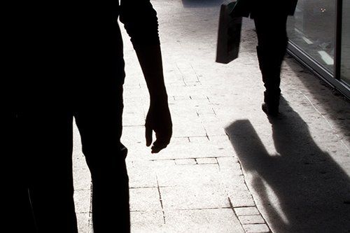 Silhouetted figures walking on a sunlit tiled sidewalk, one carrying a bag.