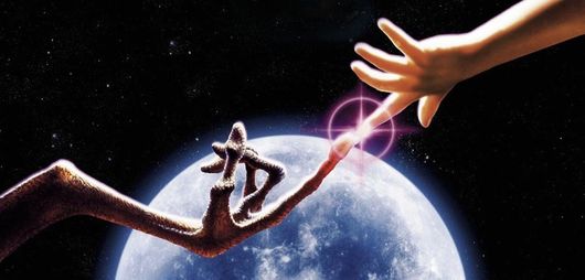 E.T. and a human hand touching fingertips, with a glowing light against a moon and starry sky.
