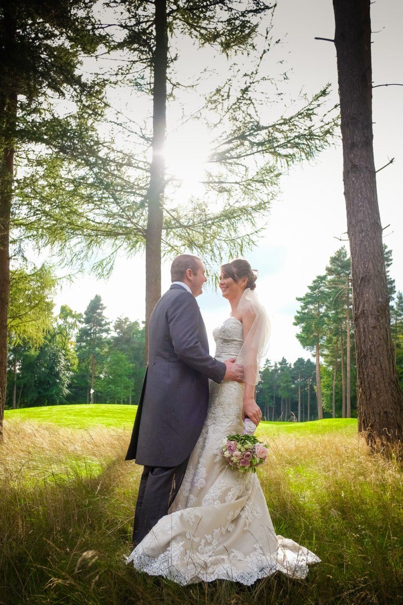 Couple in formal wear embrace, surrounded by trees and sunlight in a grassy field.