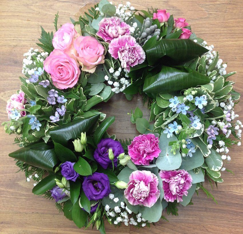Floral wreath with pink roses, carnations, and purple flowers; green foliage on wood.