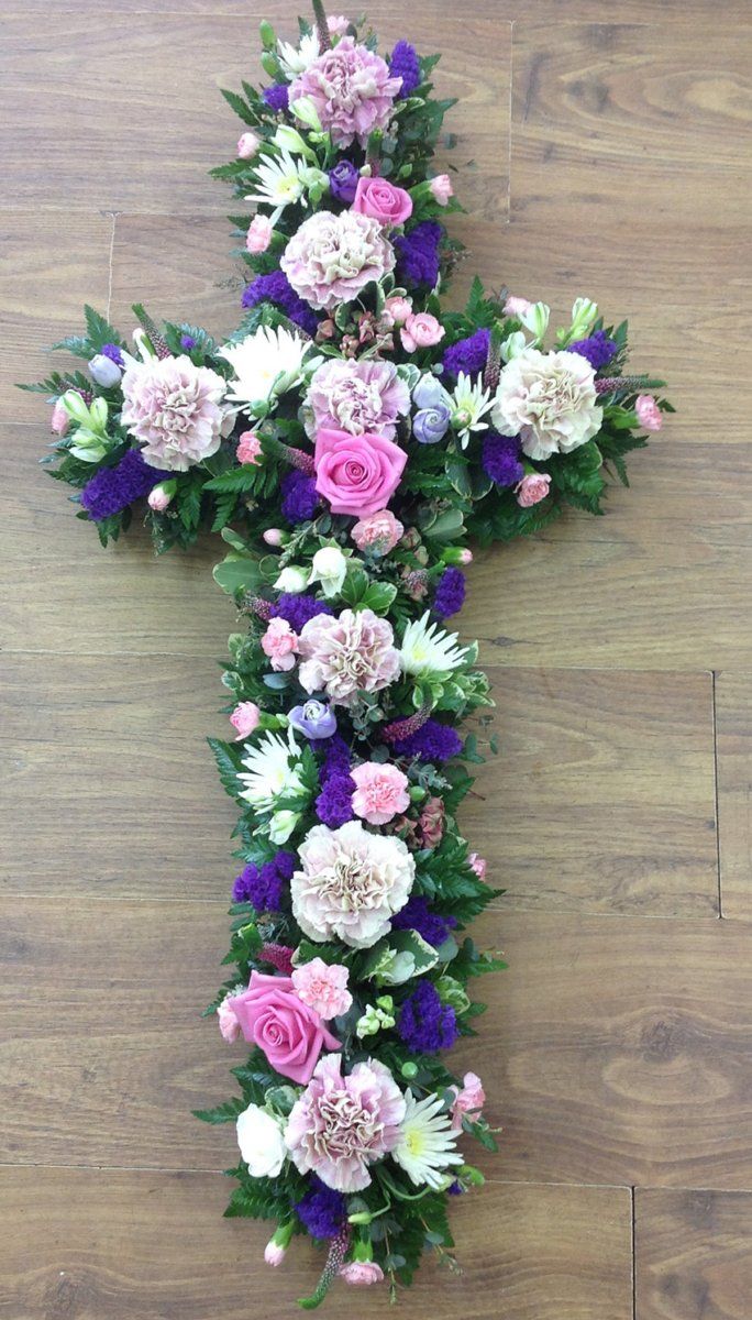 Floral cross arrangement with pink, purple, and white flowers, green foliage on a wooden surface.