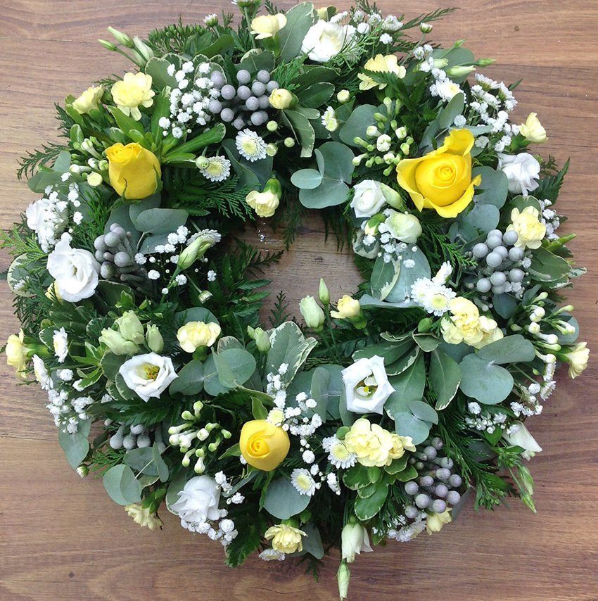 Floral wreath with yellow roses, white flowers, and green foliage on a wooden surface.