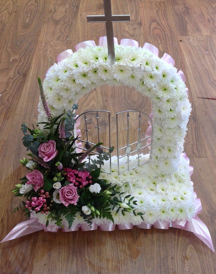 Floral arrangement, a gate of white flowers with a cross, pink roses, set on a wood floor.