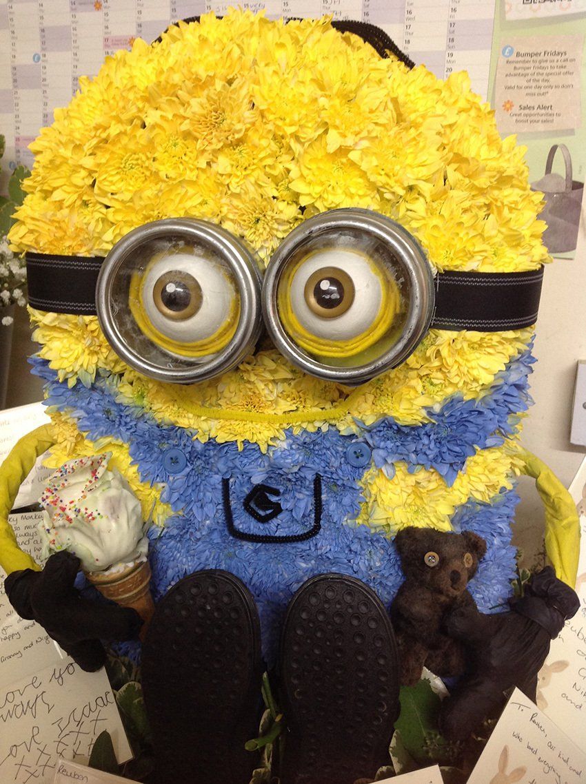 Floral arrangement of a Minion character, yellow body with blue overalls, holding a teddy bear.