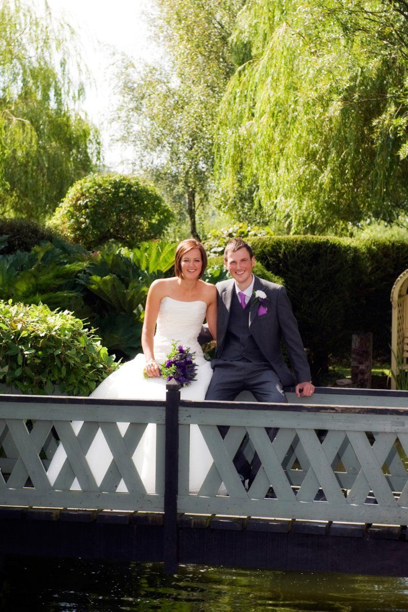 Bride and groom on a bridge. She in a white dress, he in a suit, both smiling in a garden setting.