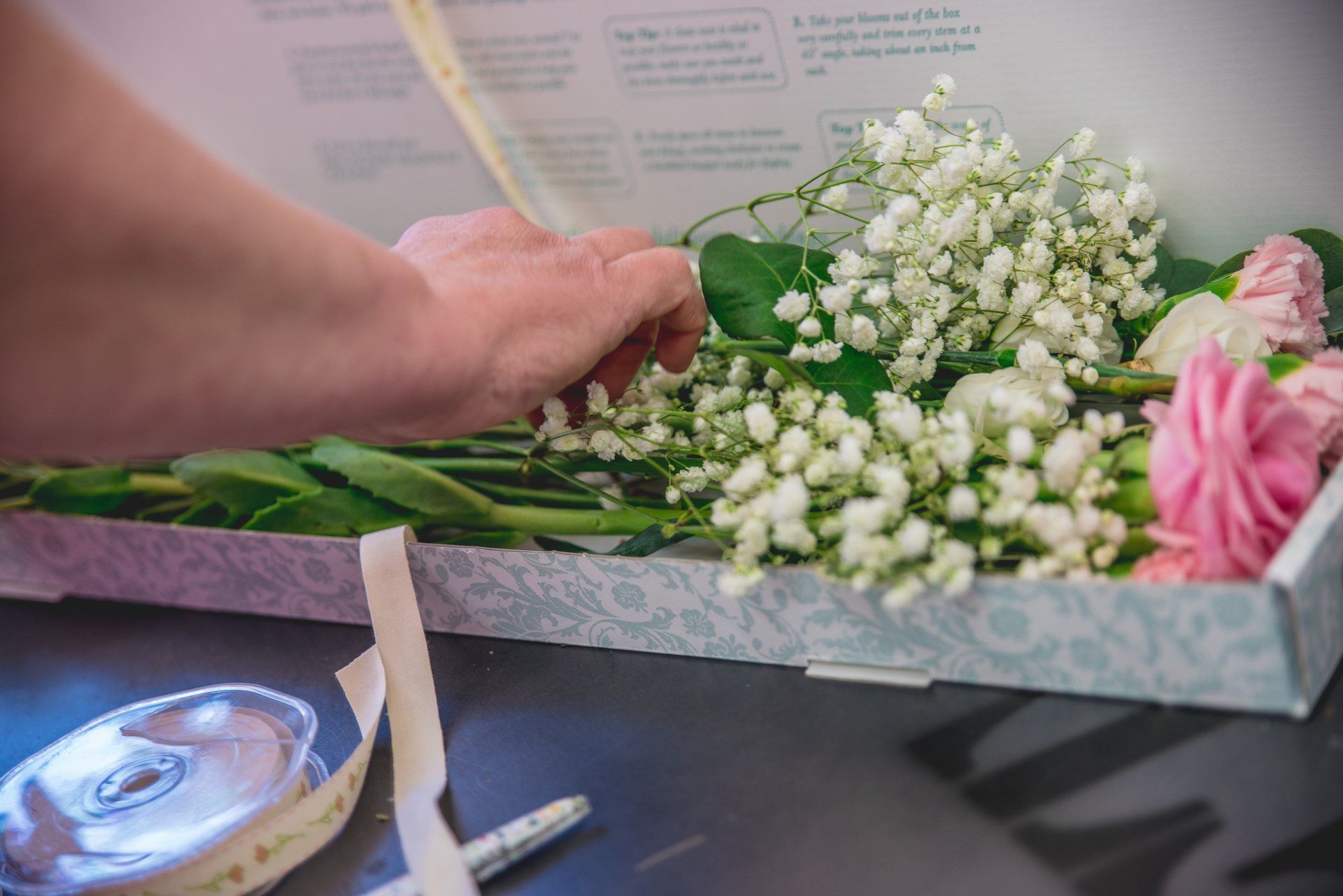 Hand arranging flowers in a box, including white and pink blossoms. Ribbon and scissors on the table.