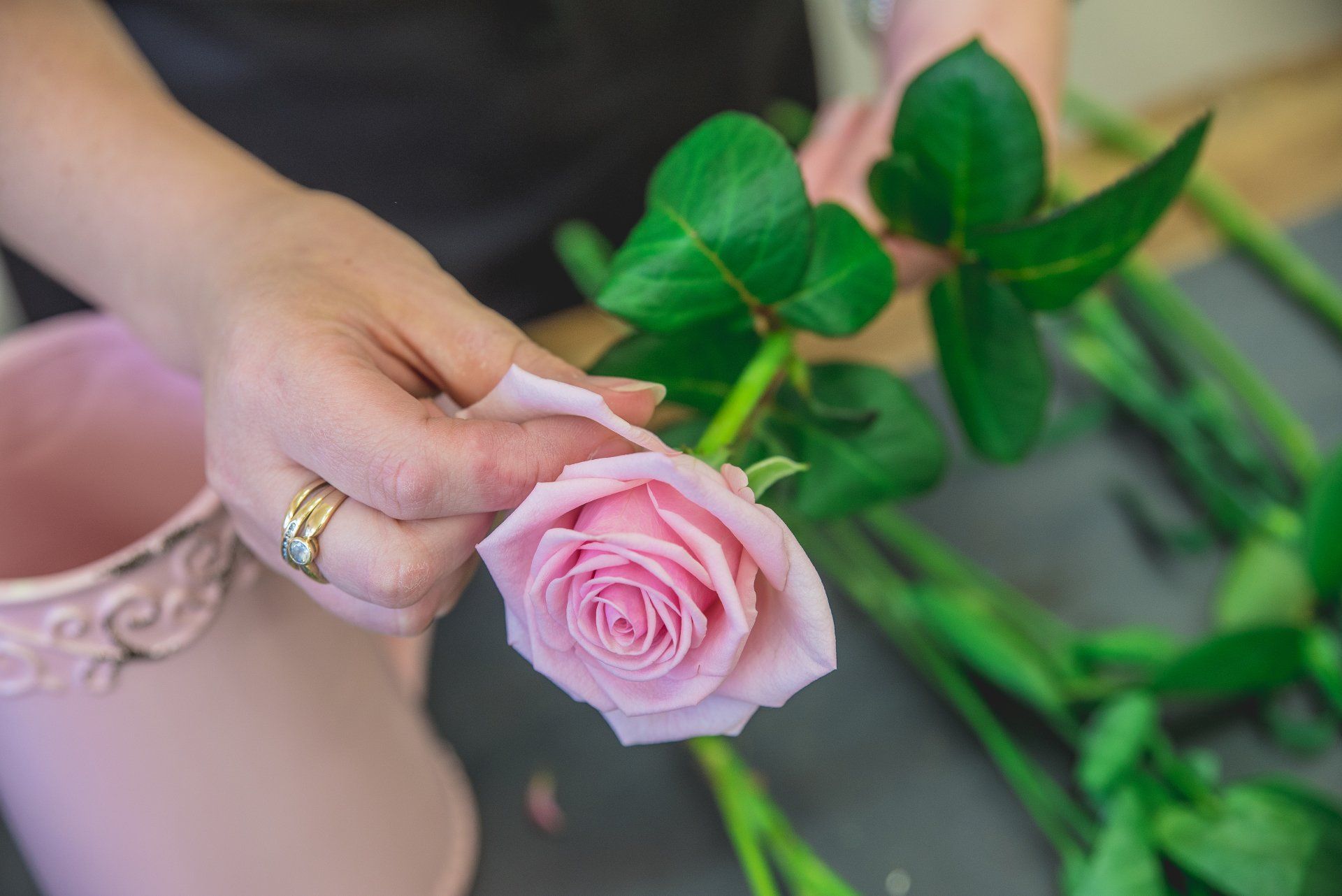 Person holding a pink rose, preparing it for an arrangement, near a pink vase and other green-stemmed flowers.