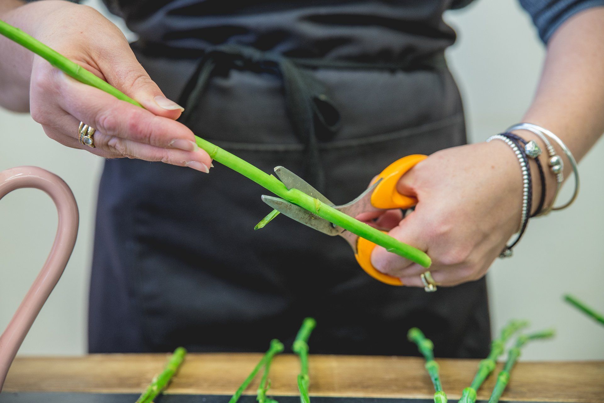Person in black apron cutting green stems with orange-handled scissors.