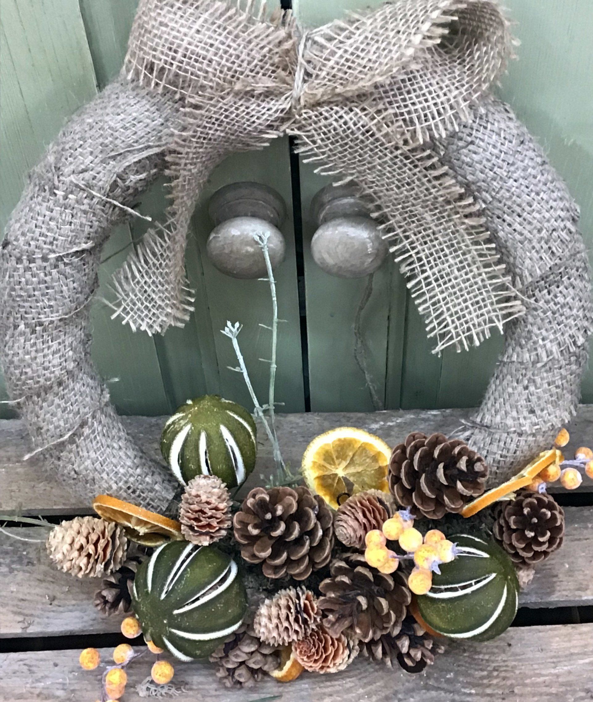 Burlap wreath decorated with pinecones, dried citrus, and green ornaments. Bow at top, resting on a wooden door.