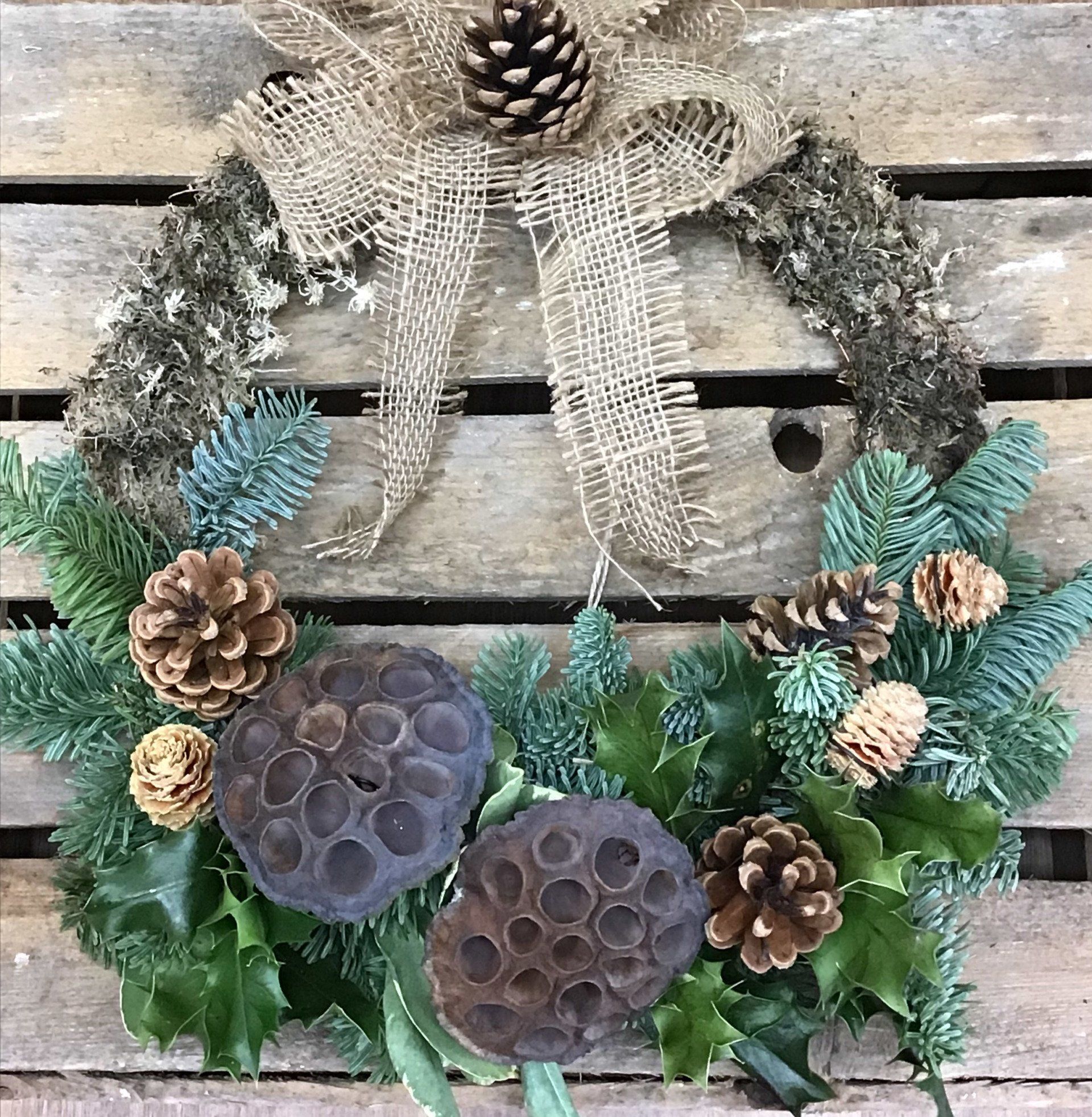 Holiday wreath with pinecones, burlap bow, greenery, and seed pods on a wood background.