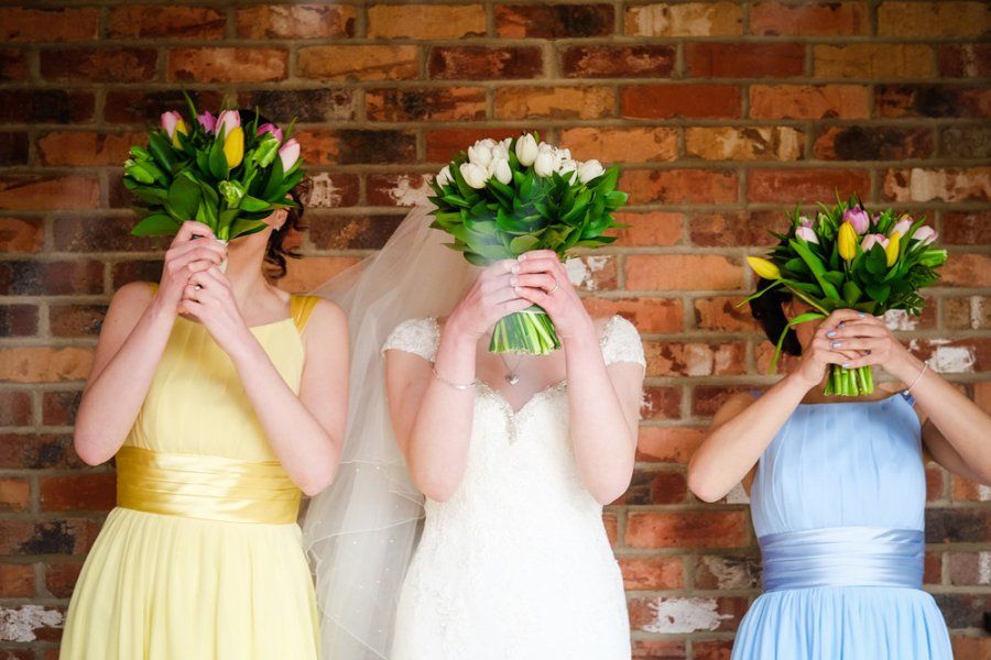 Bride and bridesmaids hold bouquets in front of their faces, standing before a brick wall.