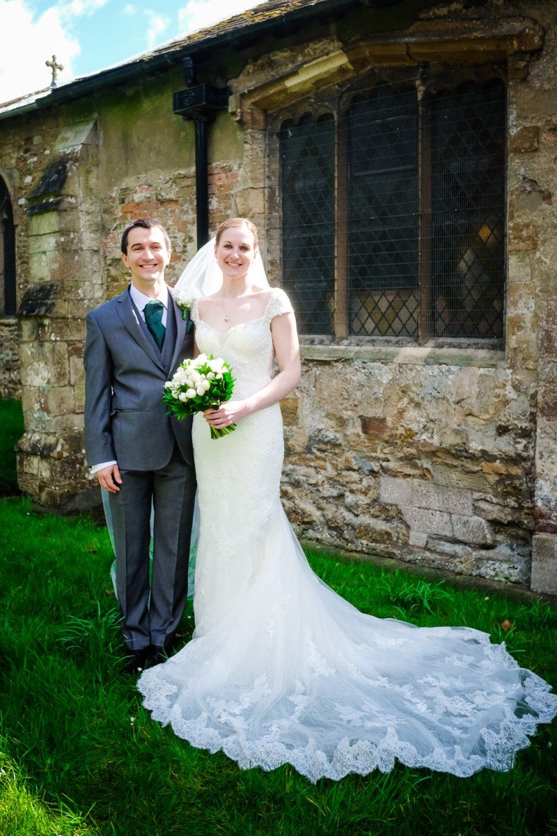 Newlyweds pose outside a stone church: groom in gray suit with teal tie, bride in white lace gown holding a bouquet.