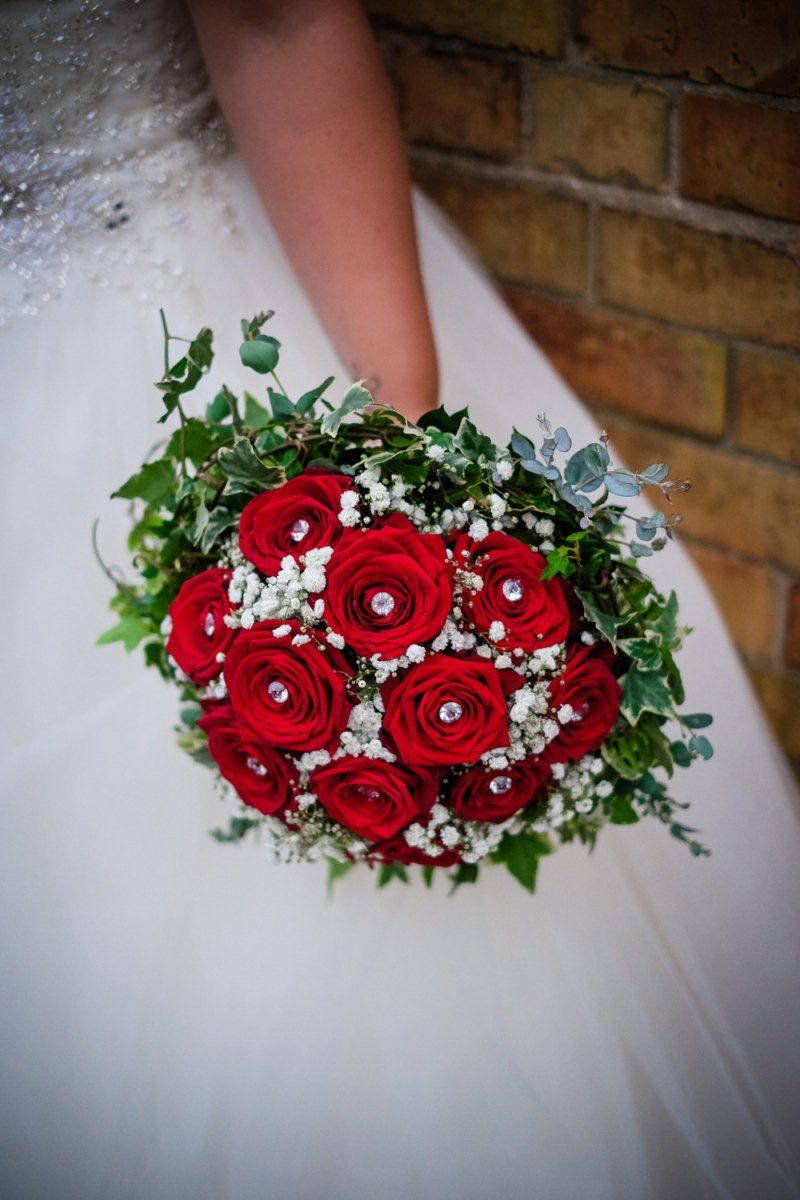 Bride holding a round bouquet of red roses with sparkling embellishments, surrounded by greenery and baby's breath.