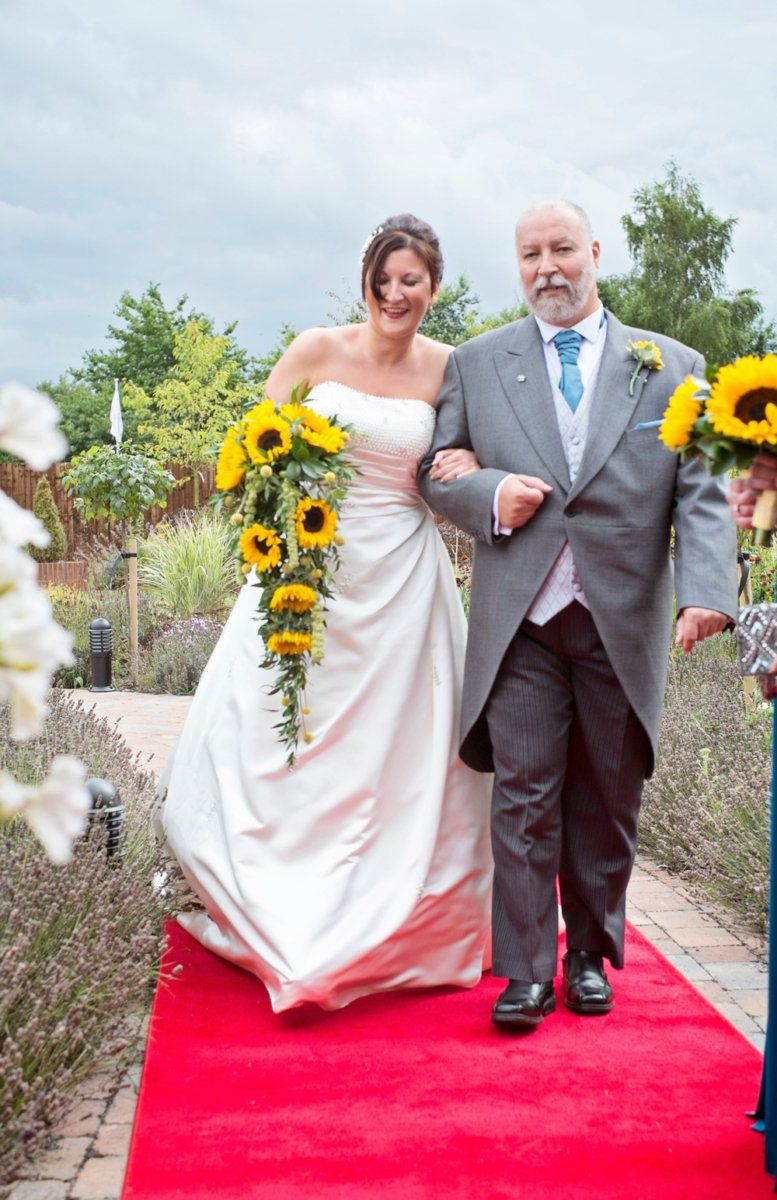 Bride in white dress holding sunflower bouquet, walks down red carpet with man in suit.