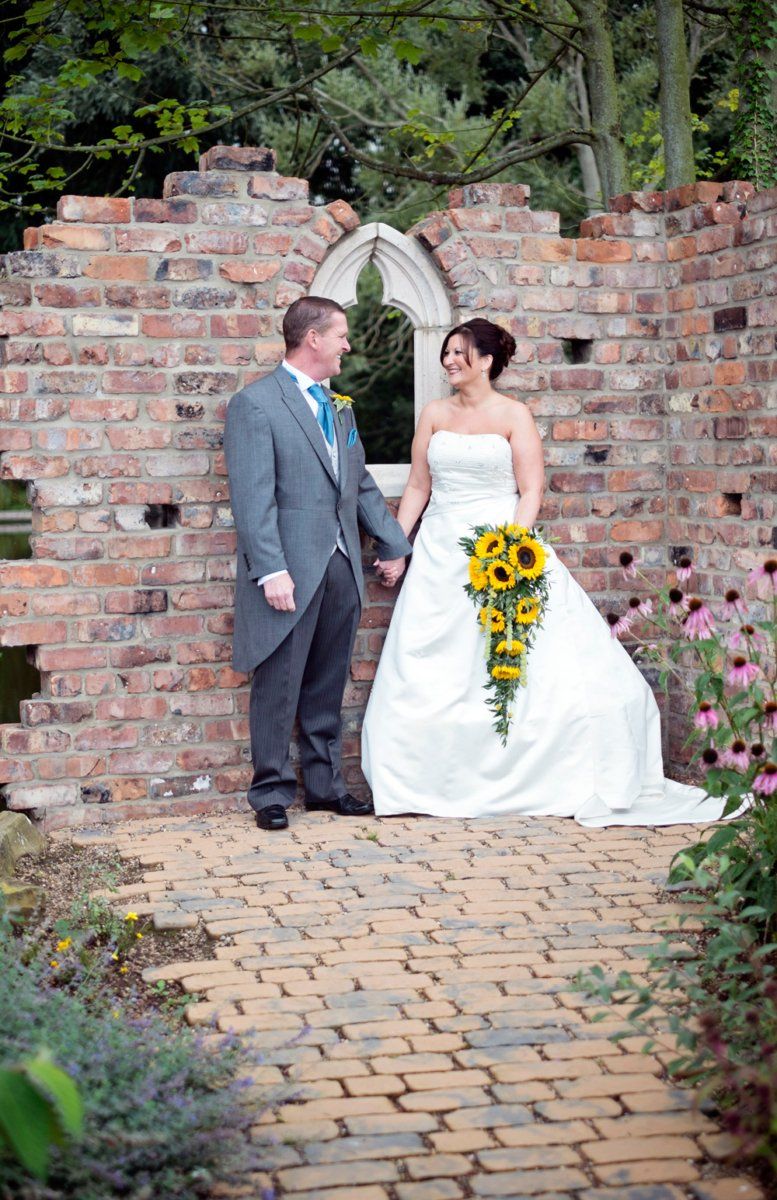 Newlyweds holding hands in front of a brick wall ruin with a sunflower bouquet.