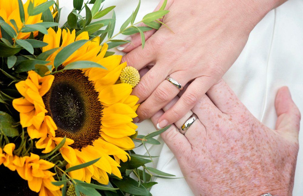 Hands with wedding rings on a white dress, near a sunflower bouquet.
