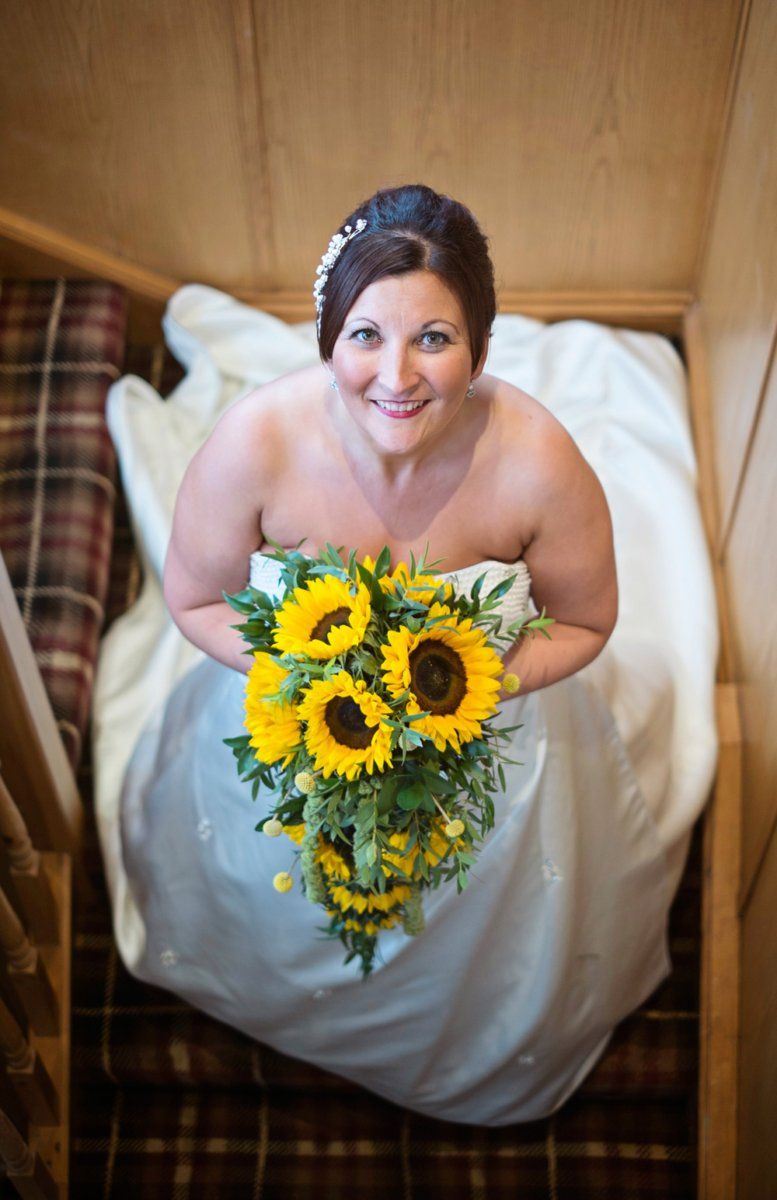 Bride in a white strapless gown holding a bouquet of sunflowers, looking up, standing on wooden stairs.