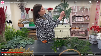 Woman arranging greenery at a table, floral supplies in a craft room.